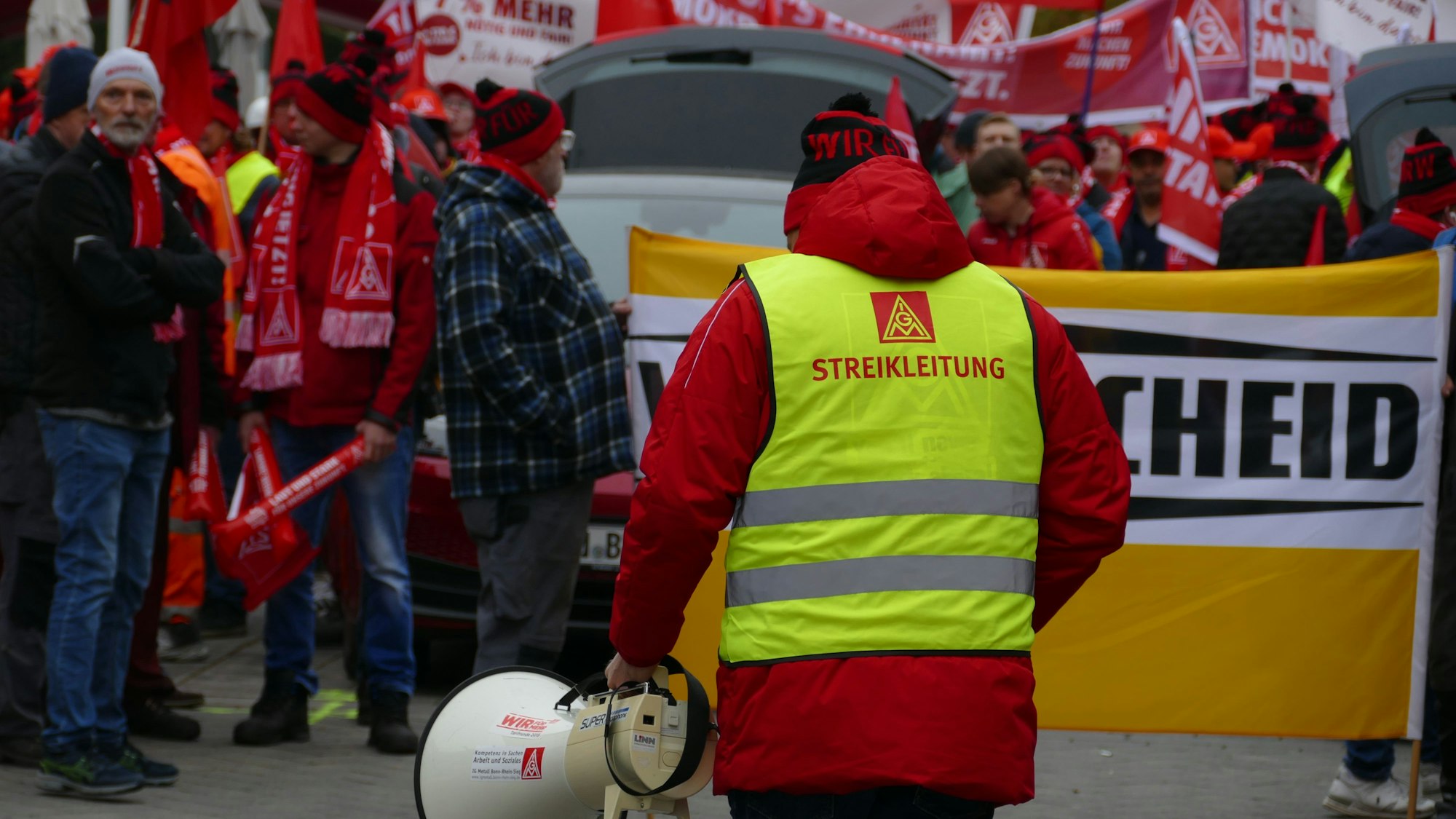Ein Mann in einer Warnweste und mit einem Megafon vor einer Menschenmenge.