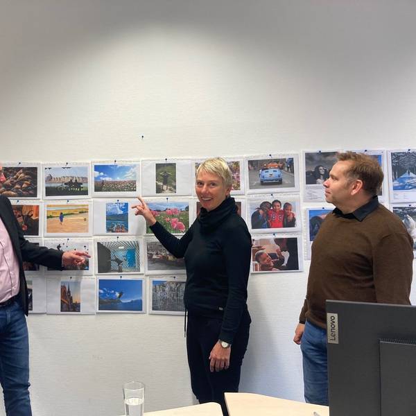 Zu sehen sind zwei Männer und eine Frau vor einer Wand mit Fotos.