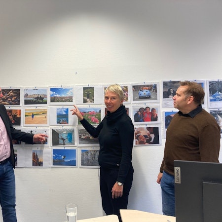 Zu sehen sind zwei Männer und eine Frau vor einer Wand mit Fotos.