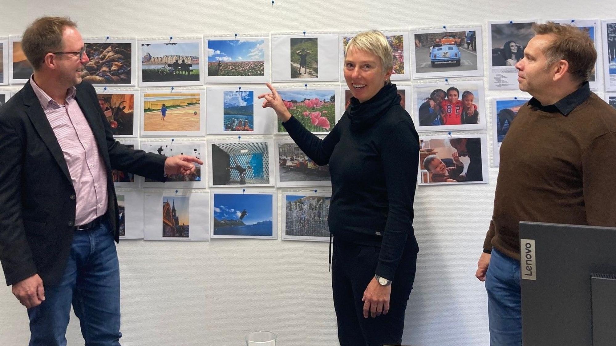 Zu sehen sind zwei Männer und eine Frau vor einer Wand mit Fotos.