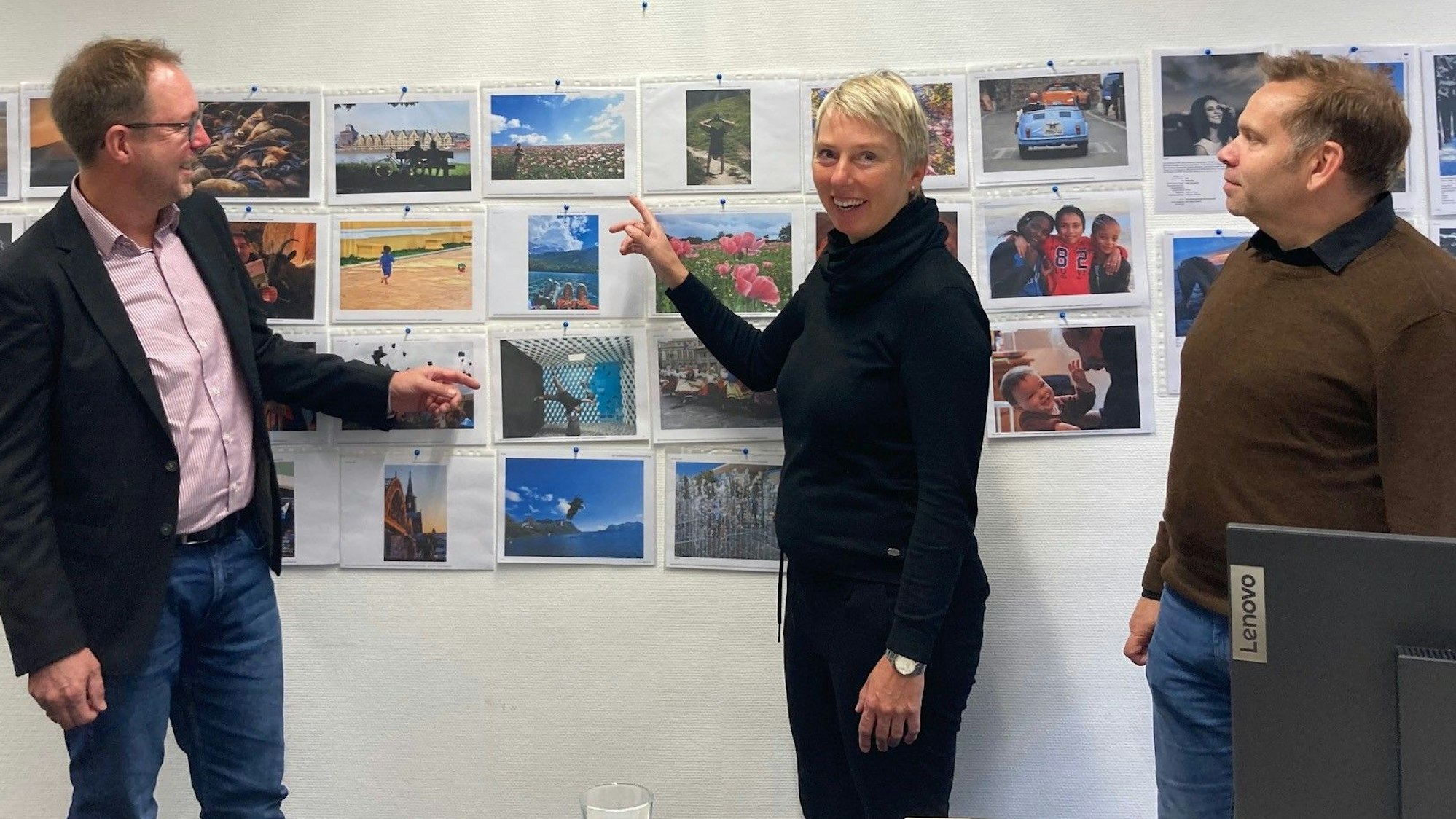 Zu sehen sind zwei Männer und eine Frau vor einer Wand mit Fotos.