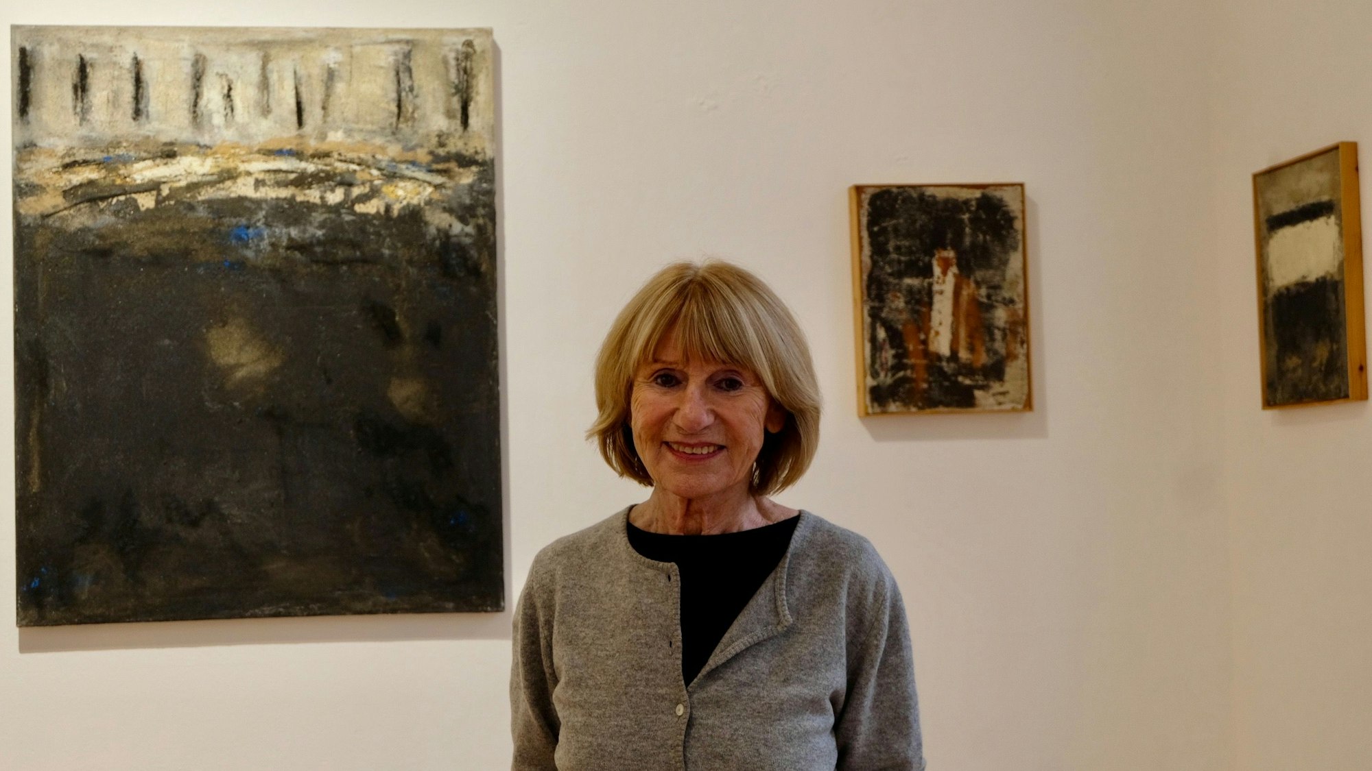 Eine Frau mit kinnlangem Haar steht vor einer Wand, an der ein großes und ein kleines Bild hängen.