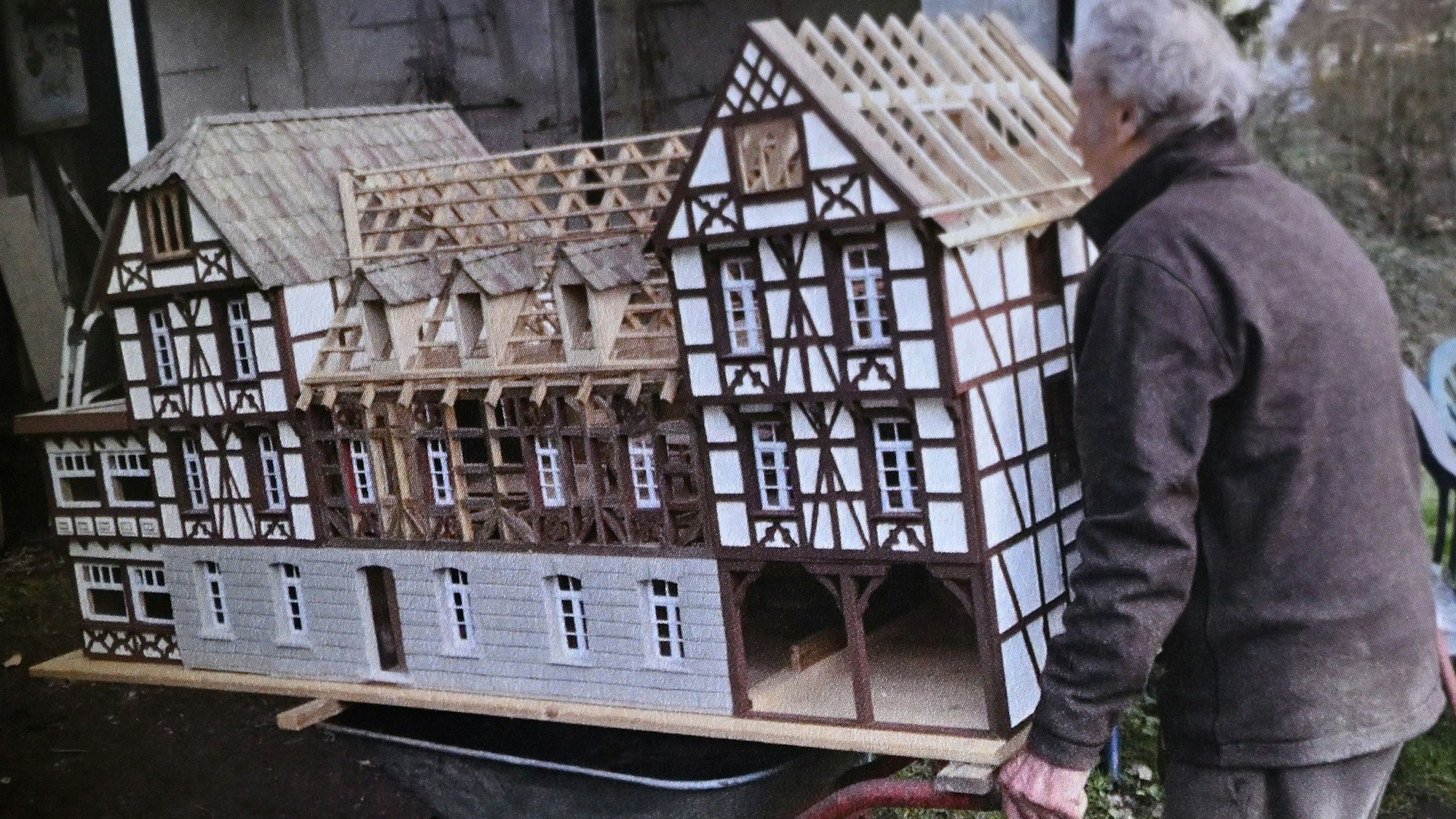 Ein alter Mann schiebt das große Modell eines Fachwerkhauses auf einer Schubkarre.