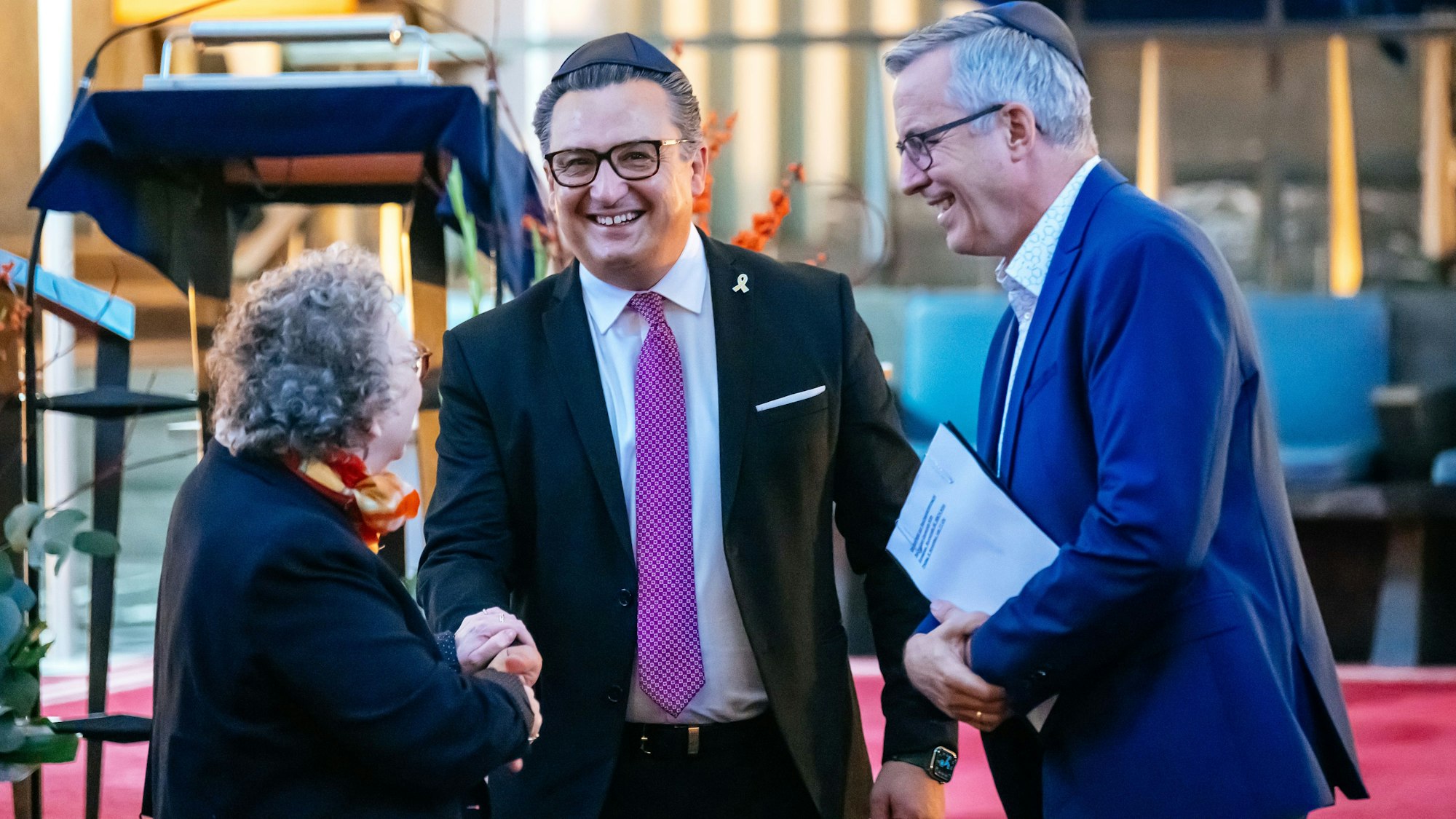 Felix Schotland, Vorstandsmitglied der Synagogen-Gemeinde Köln, und Bürgermeister Andreas Wolter (rechts) begrüßen eine Frau bei der Gedenkfeier in der Synagoge.