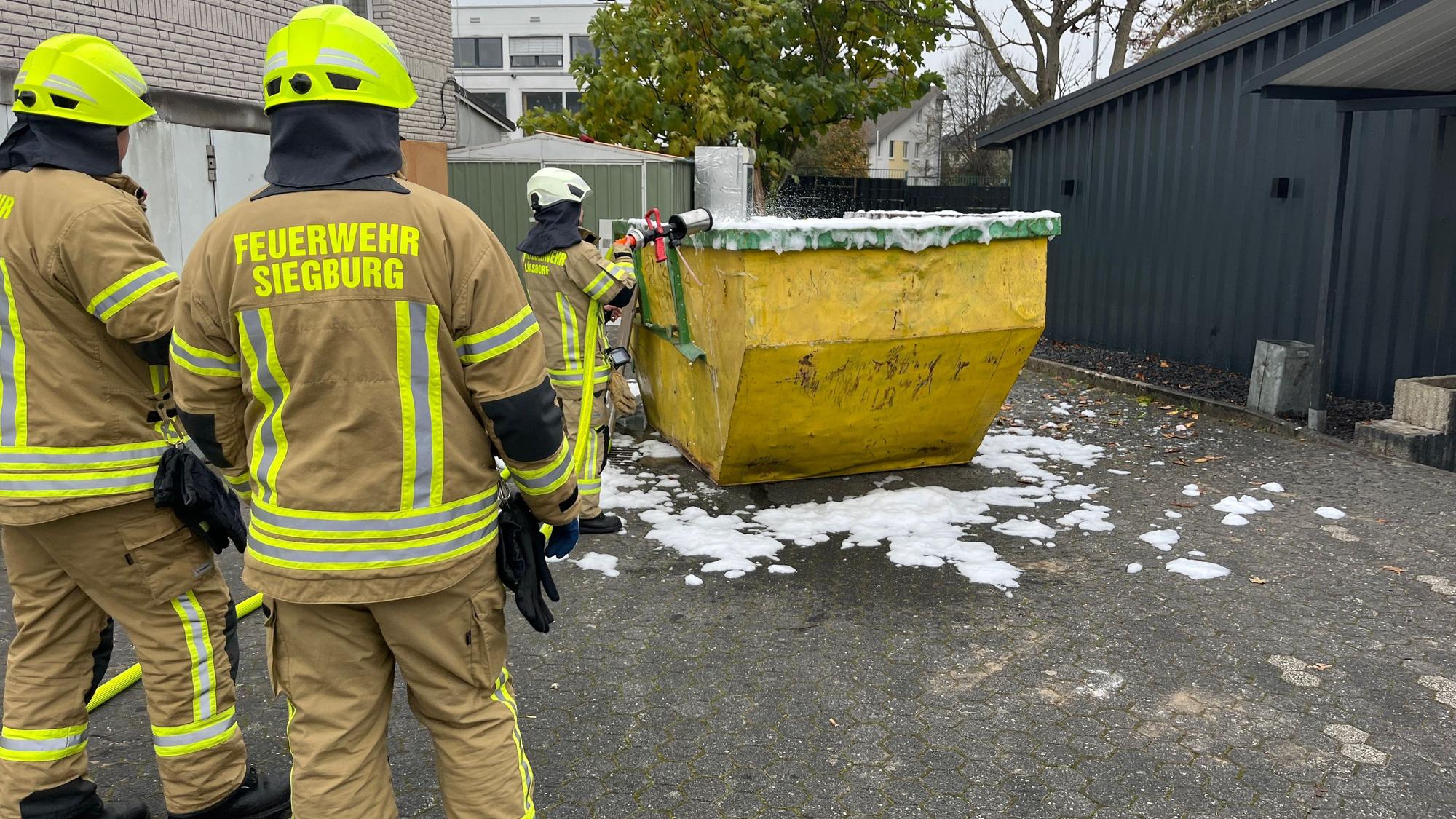 Drei Feuerwehrleute stehen vor einem Container, der mit Löschschaum geflutet wurde.