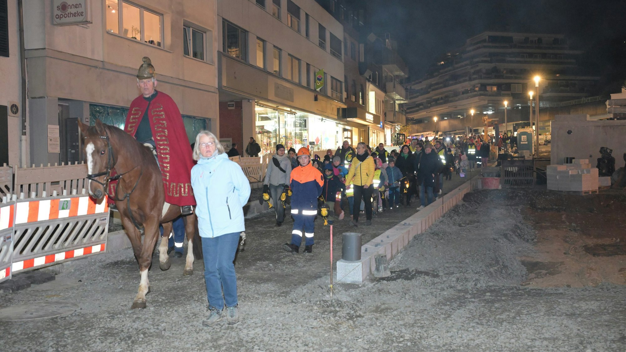 St. Martin reitet vorne weg, es folgen die Kinder mit den Laternen. Rechts und links sind die Absperrungen der Baustelle.