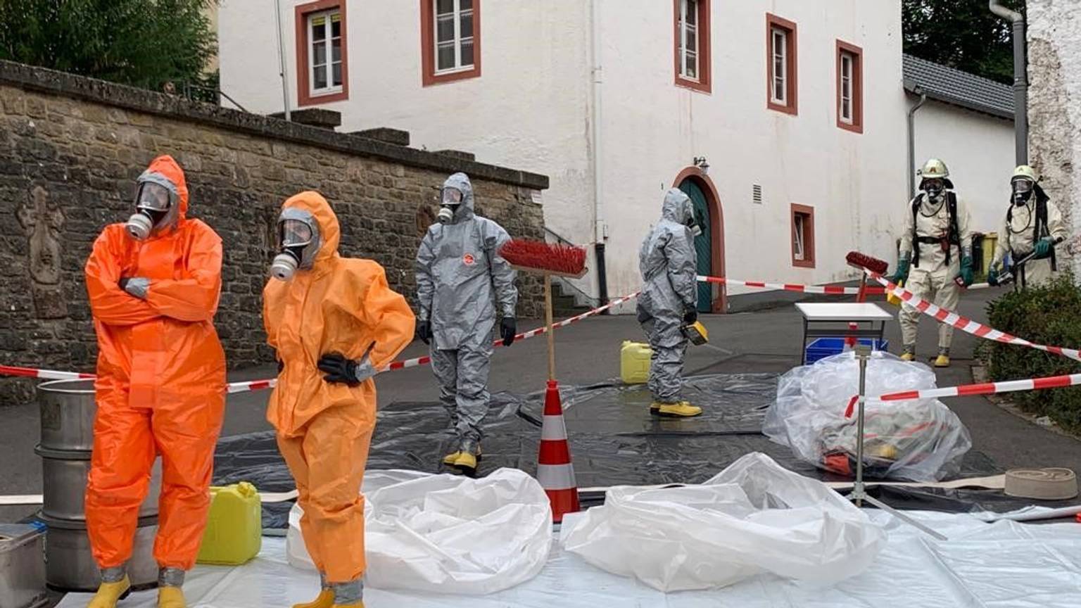 Einsatzkräfte in orangefarbenen und grauen Schutzanzügen stehen auf einer weißen Plane vor einem Haus.