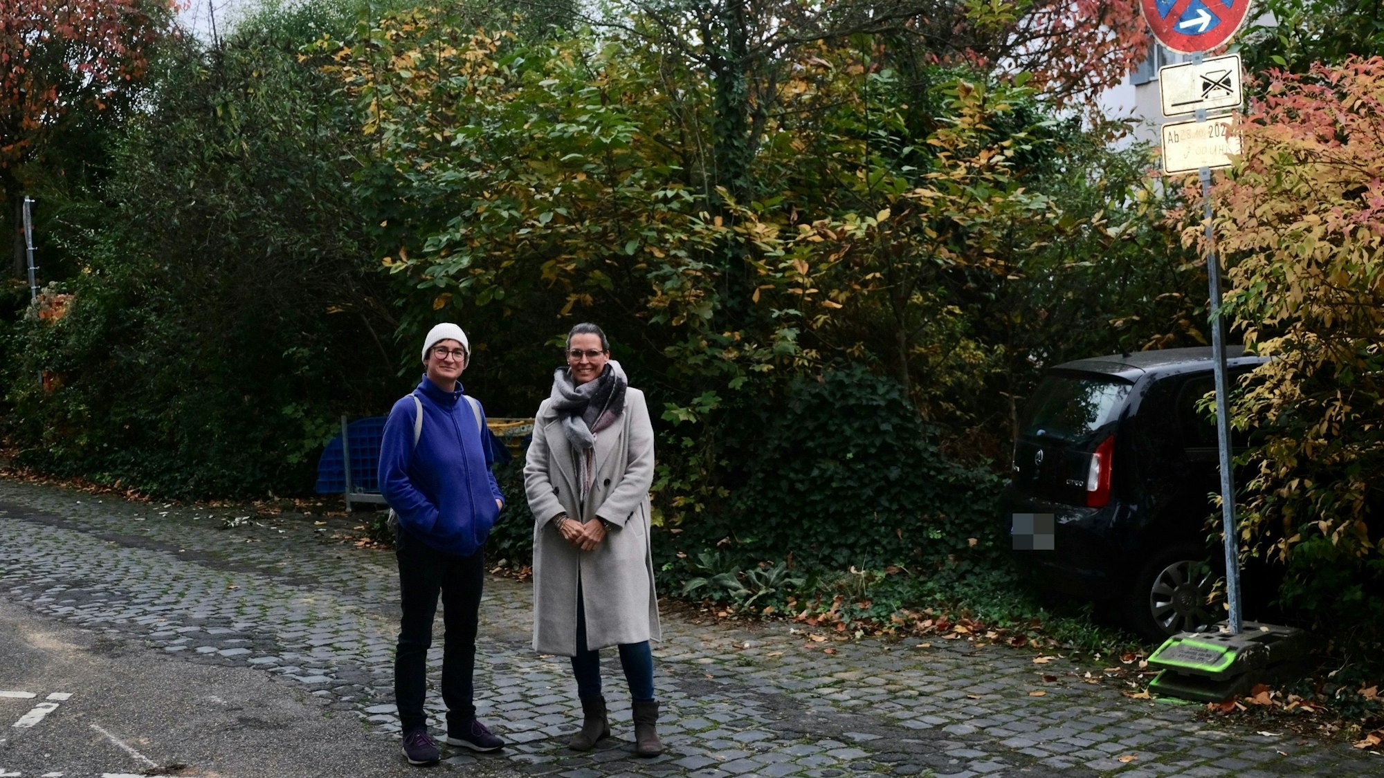 Zwei Frauen stehen auf einer Straße, hinter ihnen ist üppig wucherndes Grün zu sehen.