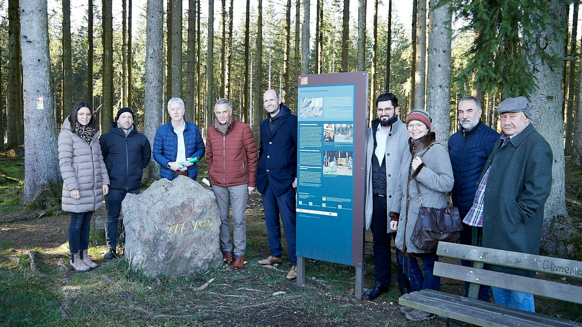 Eine Gruppe von Menschen steht neben einem Gedenkstein und einer Tafel im Wald.