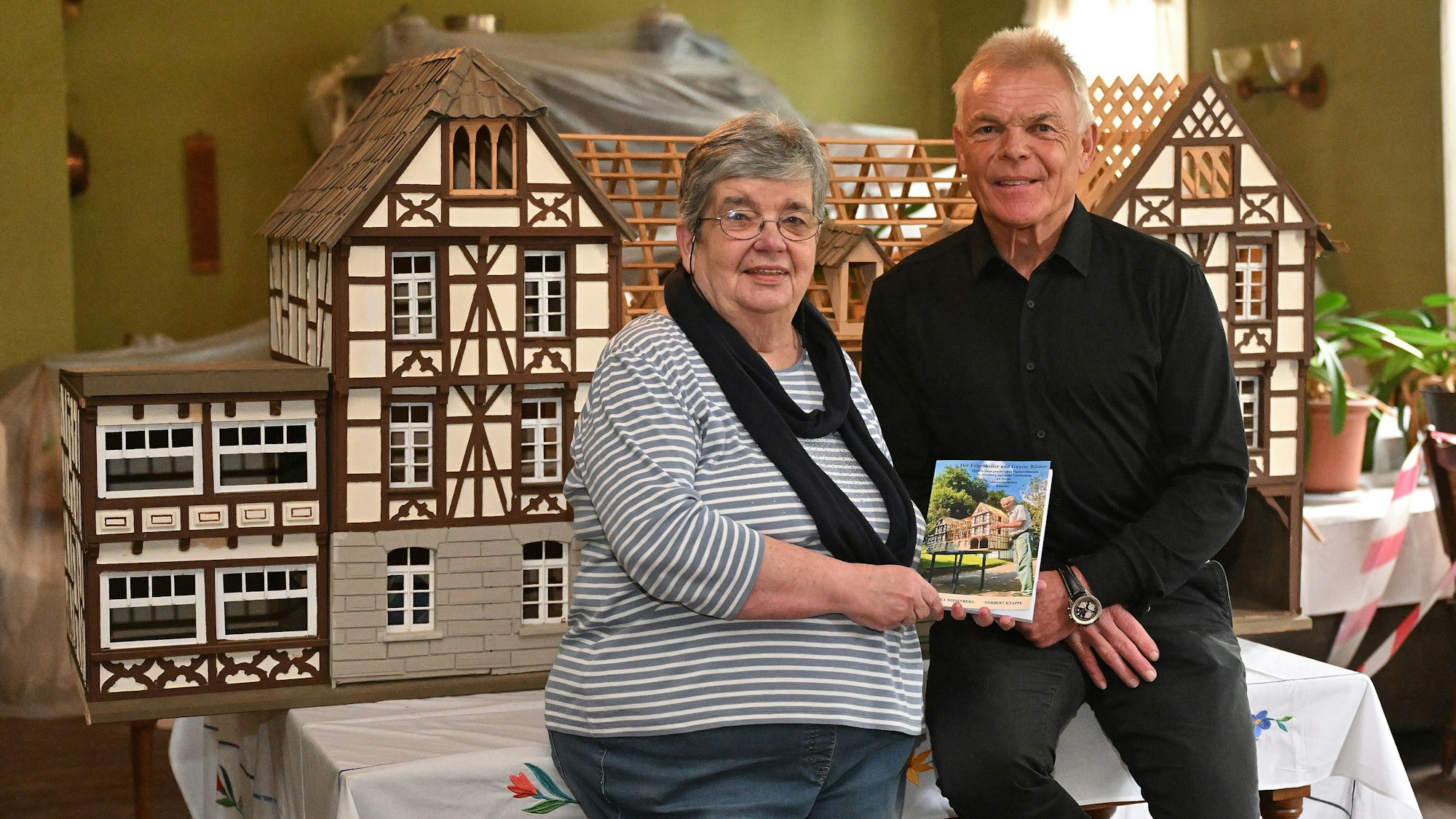 Ein großes Modell eines Fachwerkhauses auf einem Tisch, davor stehen ein Mann und eine Frau und präsentieren ein Buch.