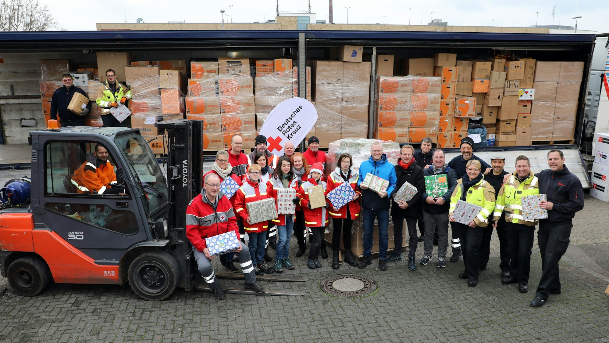 Menschen mit Paketen stehen neben einem Gabelstapler vor einem Sattelzugauflieger.