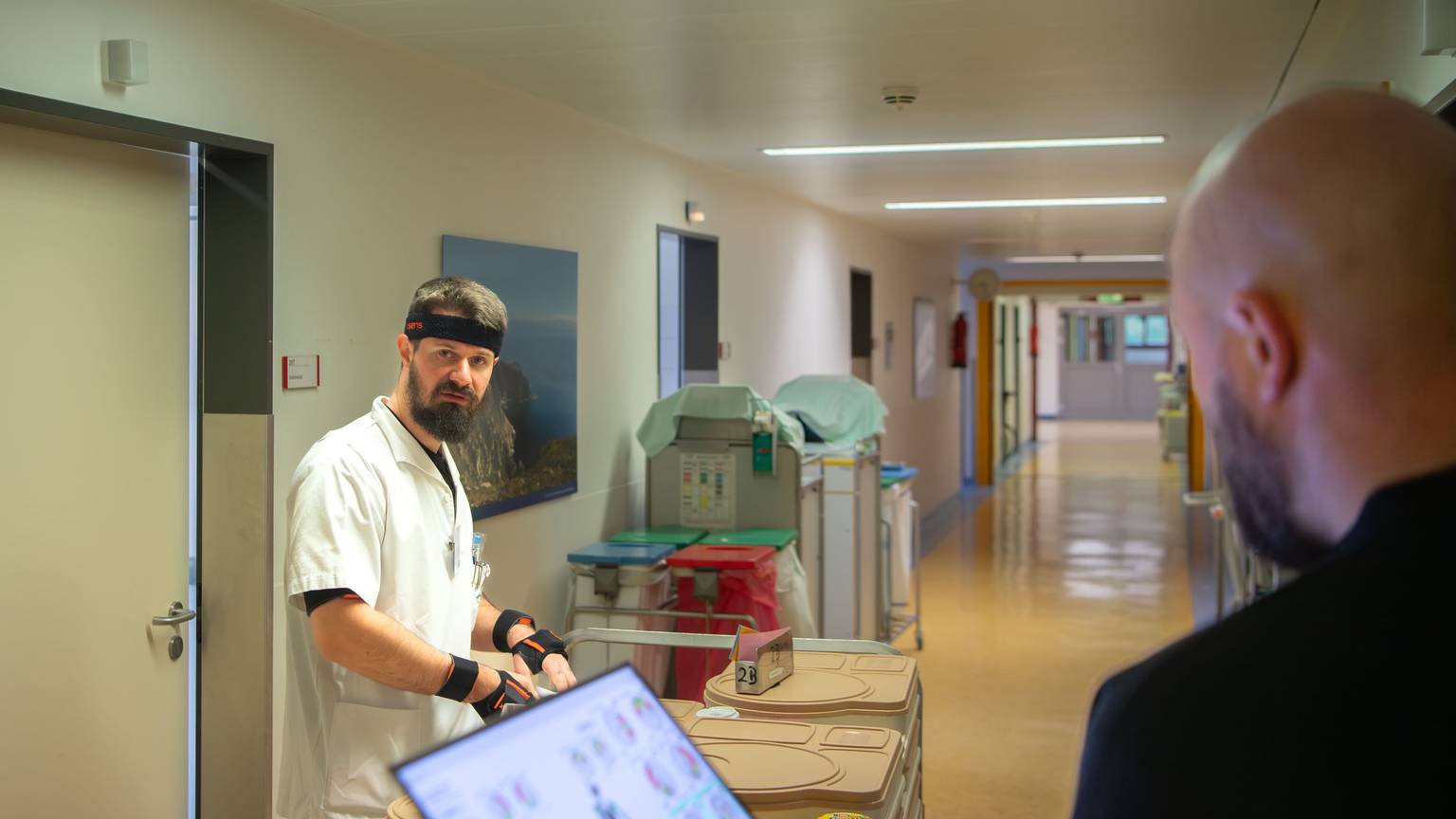 Ein Mitarbeiter in weißer Kleidung steht auf einem Krankenhausflur. An Kopf und Armen trägt er Sensoren. Ein anderer Mann an einem Laptop beobachtet ihn.
