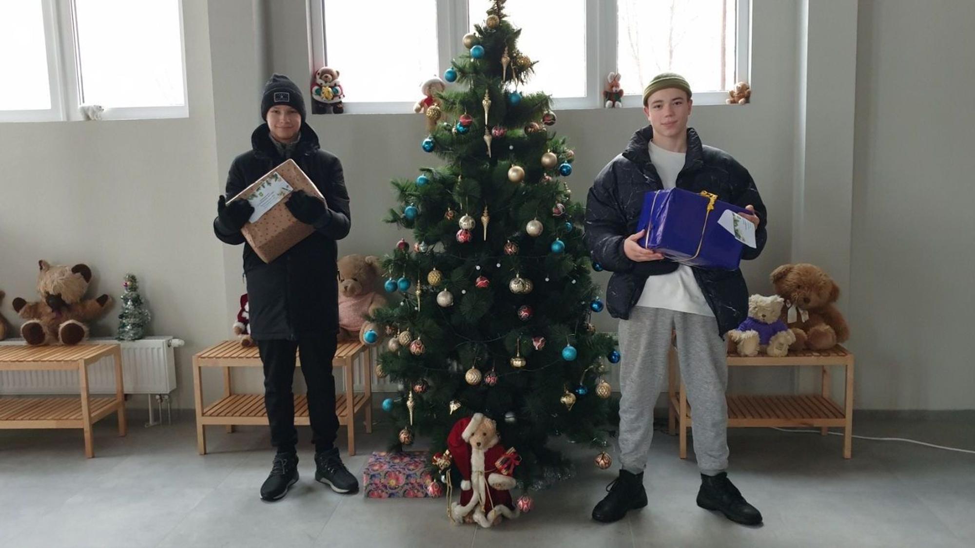 Kinder stehen mit zwei Paketen in Jacken und Mützen neben einem Weihnachtsbaum in einer Halle