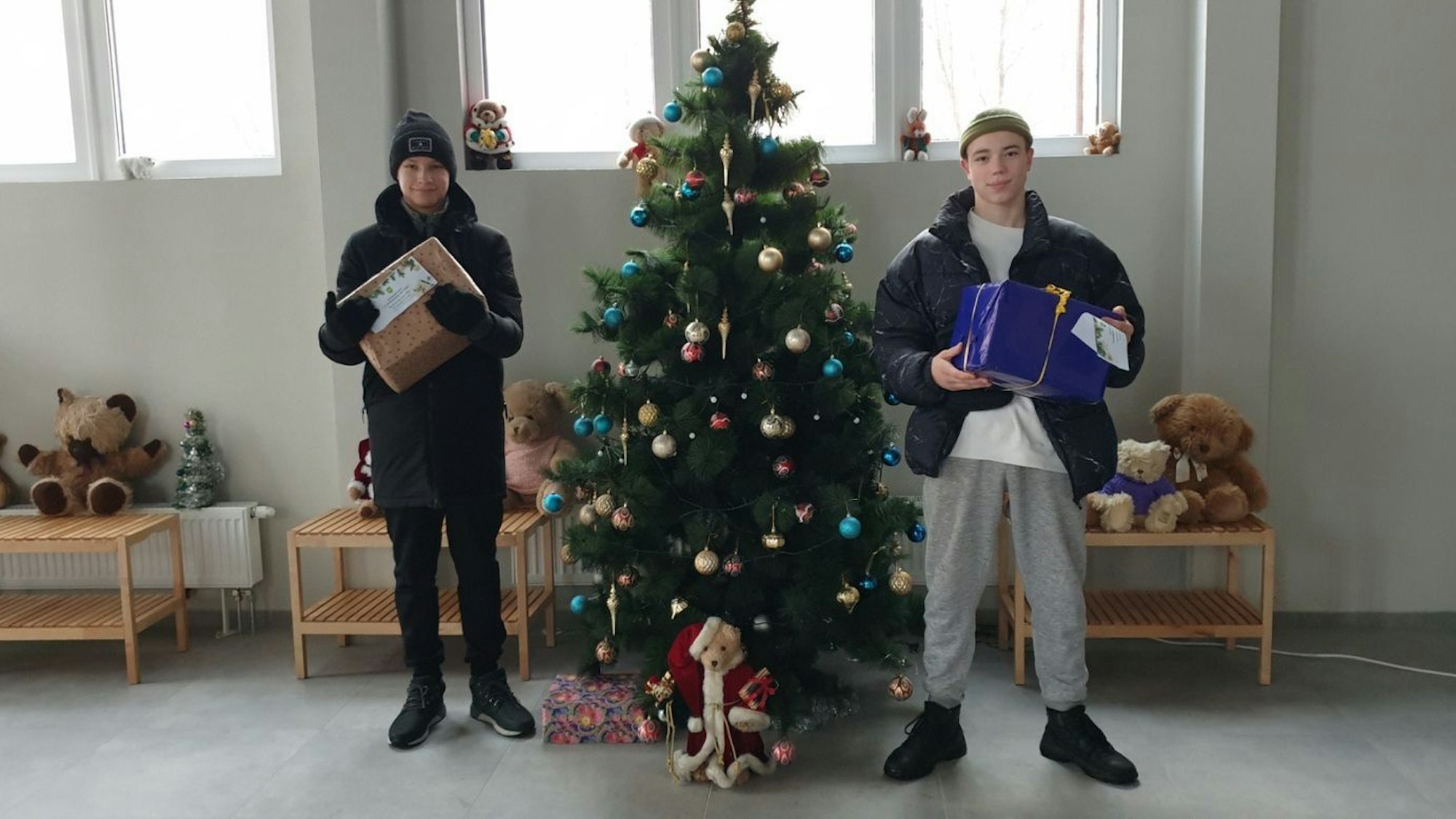 Kinder stehen mit zwei Paketen in Jacken und Mützen neben einem Weihnachtsbaum in einer Halle