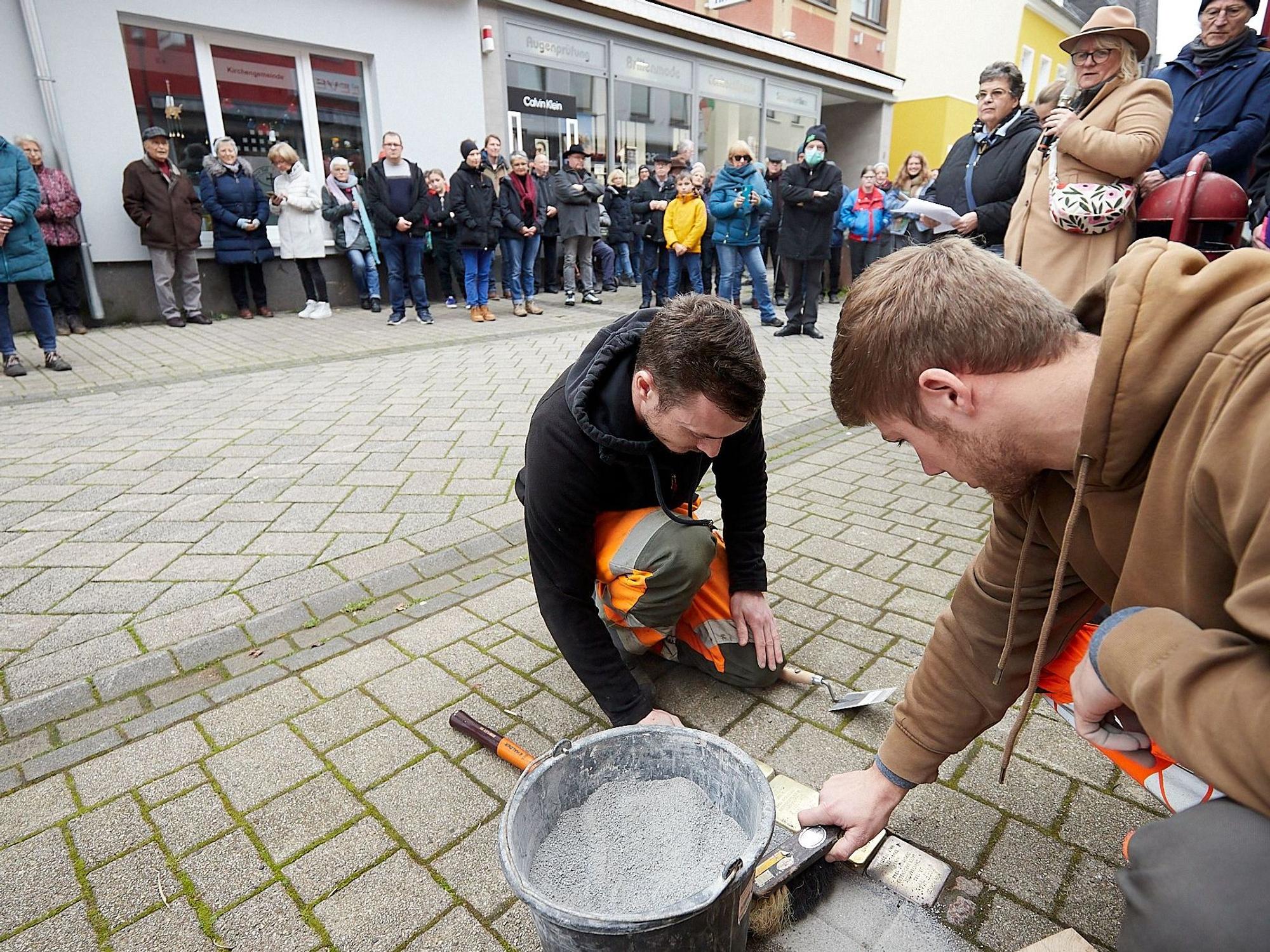Zwei Männer verlegen einige Stolpersteine auf dem Gehweg, während viele Menschen dabei zuschauen.