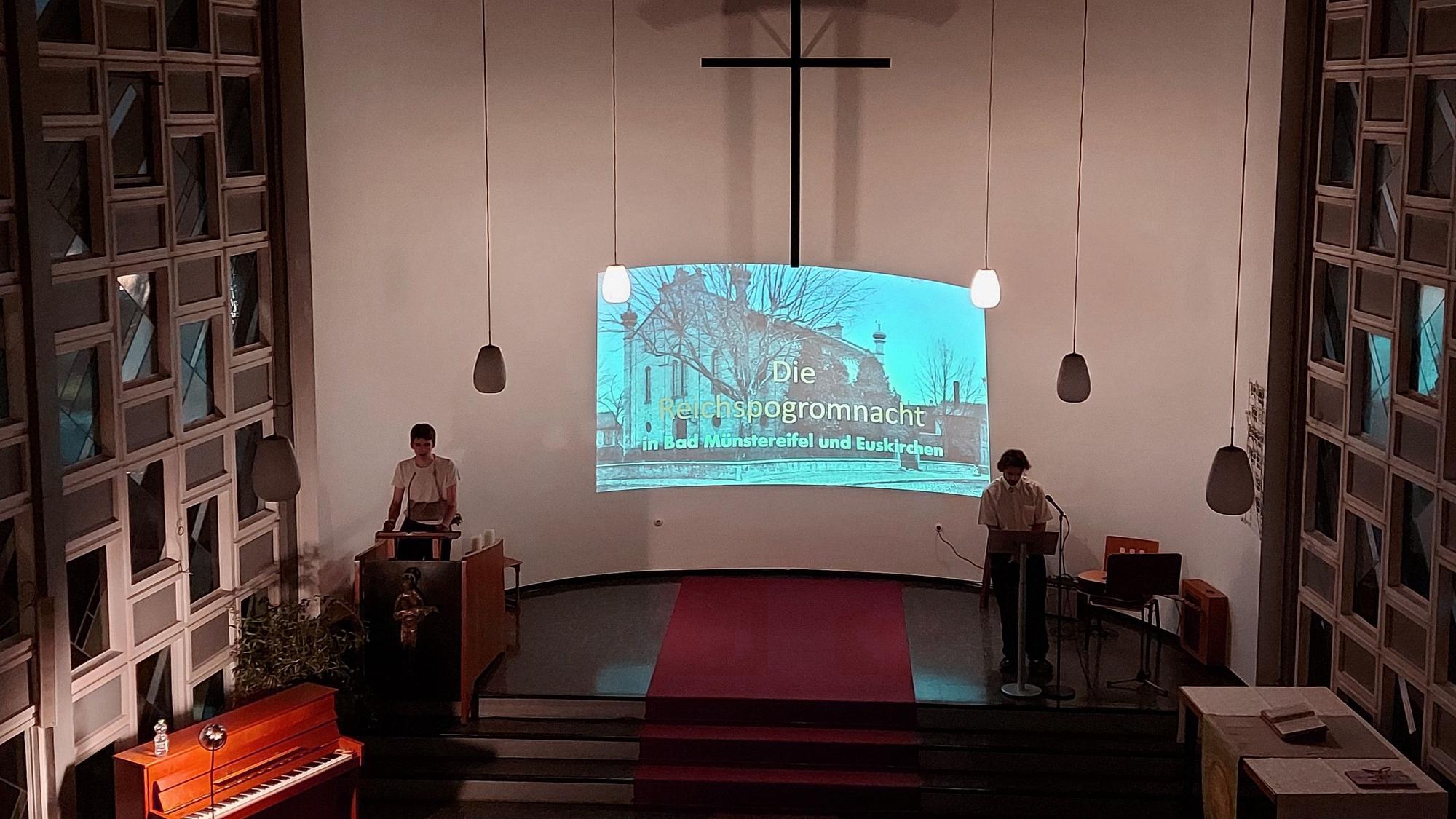 Im Altarraum einer evangelischen Kirche stehen zwei Schüler, im Hintergrund sieht man Fotos von einer Fahrt in die Gedenkstätte des ehemaligen Konzentrationslagers Auschwitz.
