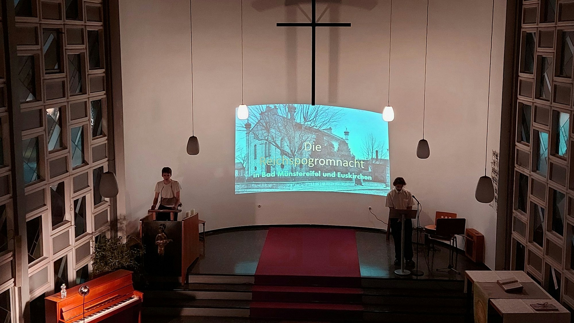 Im Altarraum einer evangelischen Kirche stehen zwei Schüler, im Hintergrund sieht man Fotos von einer Fahrt in die Gedenkstätte des ehemaligen Konzentrationslagers Auschwitz.