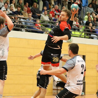 Handballer Robin Voiß steigt zum Sprungwurf hoch. Um ihn herum drei Gegenspieler, direkt vor ihm springt ein weiterer mit erhobenen Armen hoch. Im Hintergrund ist die Zuschauertribüne zu erkennen.