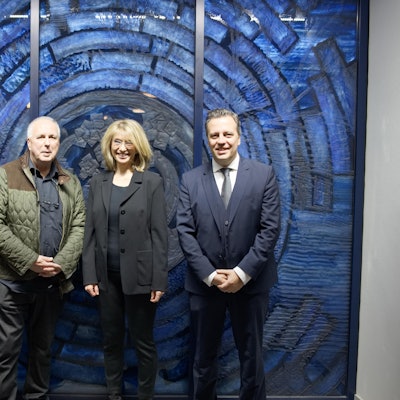 Eine Gruppe von drei Männern und einer Frau stehen vor einem wandgroßen, blauen Glaskunstwerk.