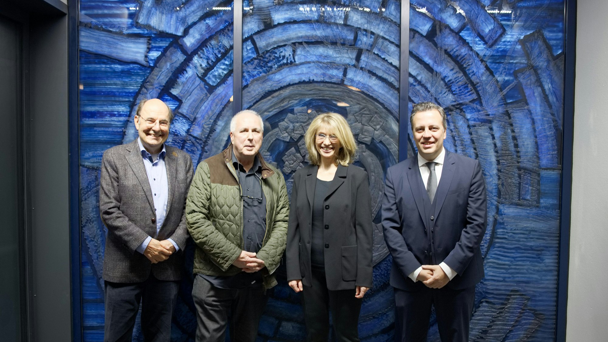 Eine Gruppe von drei Männern und einer Frau stehen vor einem wandgroßen, blauen Glaskunstwerk.