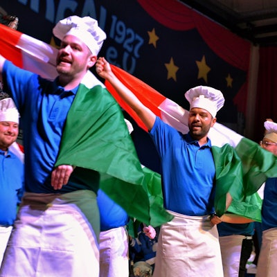 Als Köche verkleidete Männer stehen tanzend auf der Bühne und schwenken italienische Flaggen.