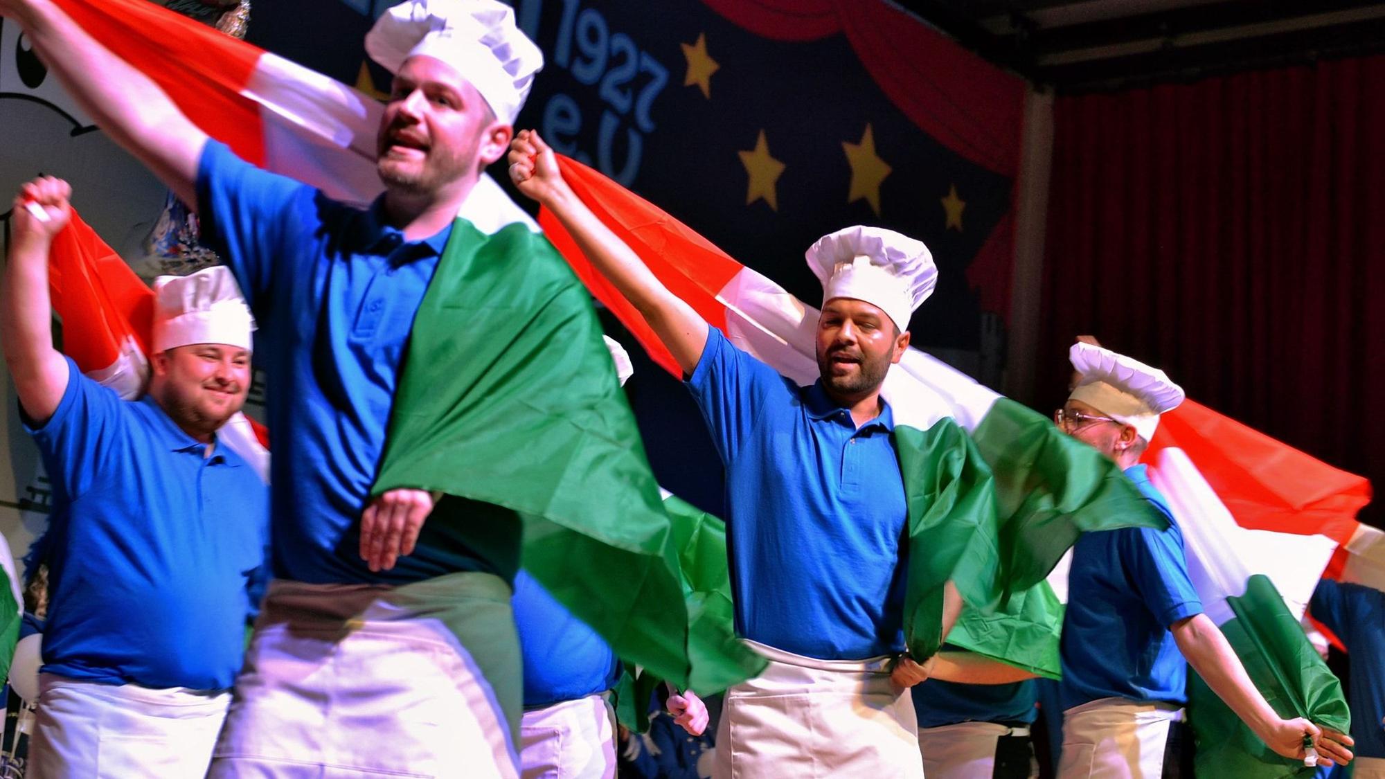 Als Köche verkleidete Männer stehen tanzend auf der Bühne und schwenken italienische Flaggen.