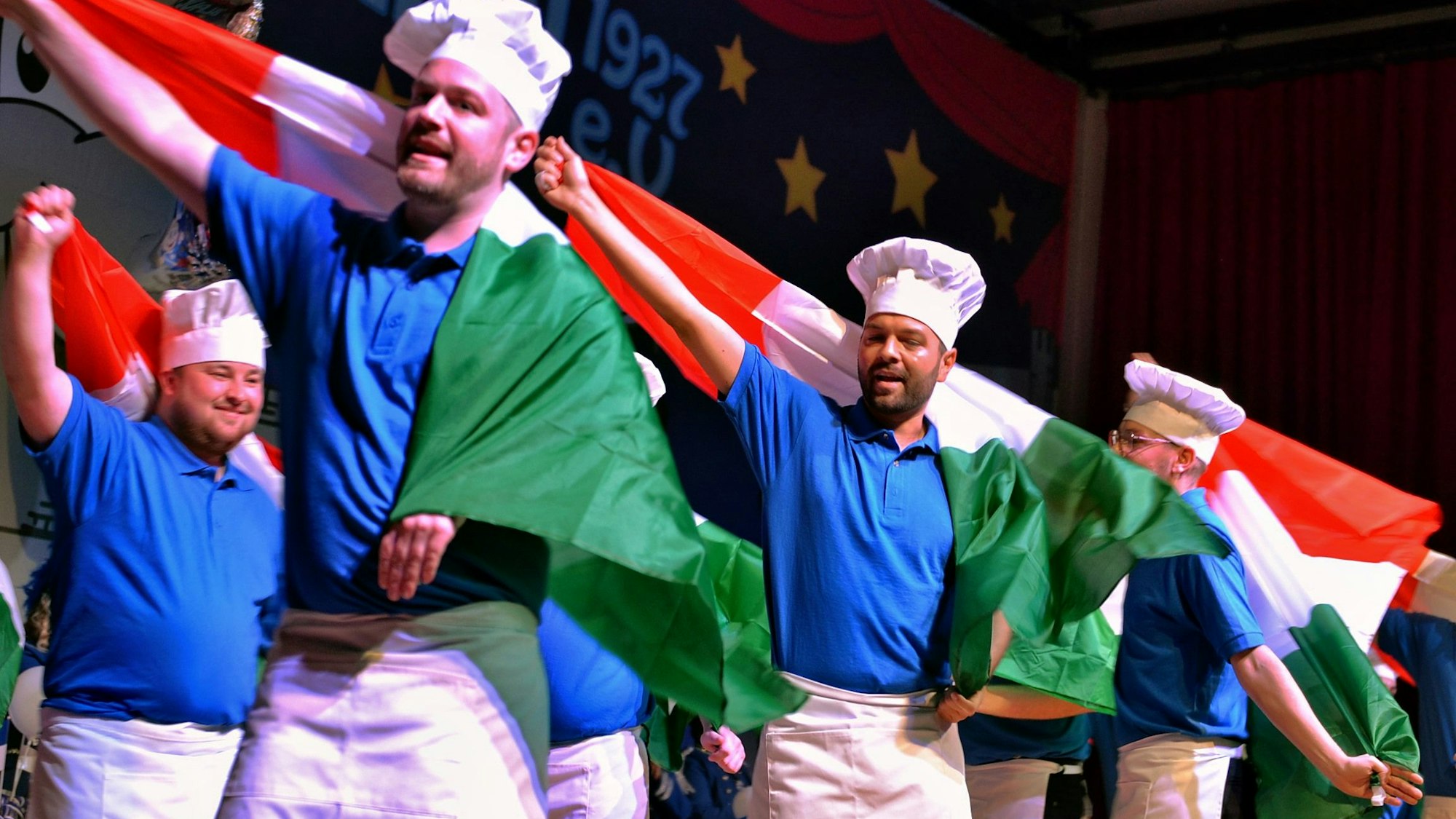 Als Köche verkleidete Männer stehen tanzend auf der Bühne und schwenken italienische Flaggen.