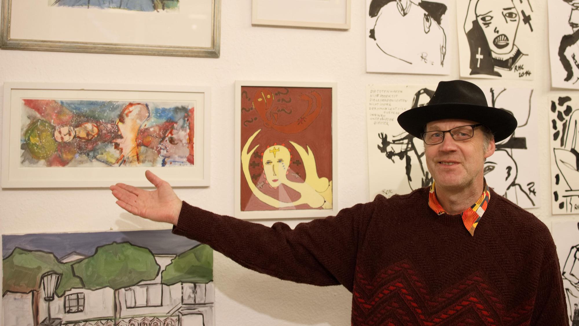 Ein Mann mit Hut zeigt auf ein gerahmtes Bild, das neben anderen Bildern an einer weißen Wand hängt.