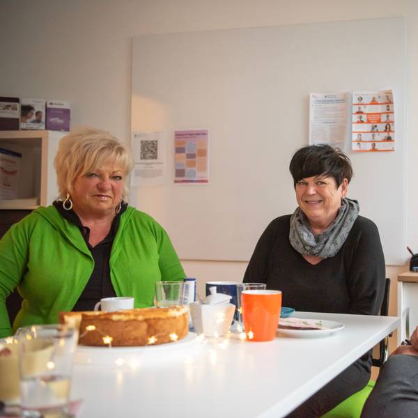 Die drei Frauen sitzen an einem Tisch in einem Büroraum. Auf dem Tisch stehen ein Käsekuchen und Kaffeegeschirr, daneben liegt eine Sternen-Lichterkette.