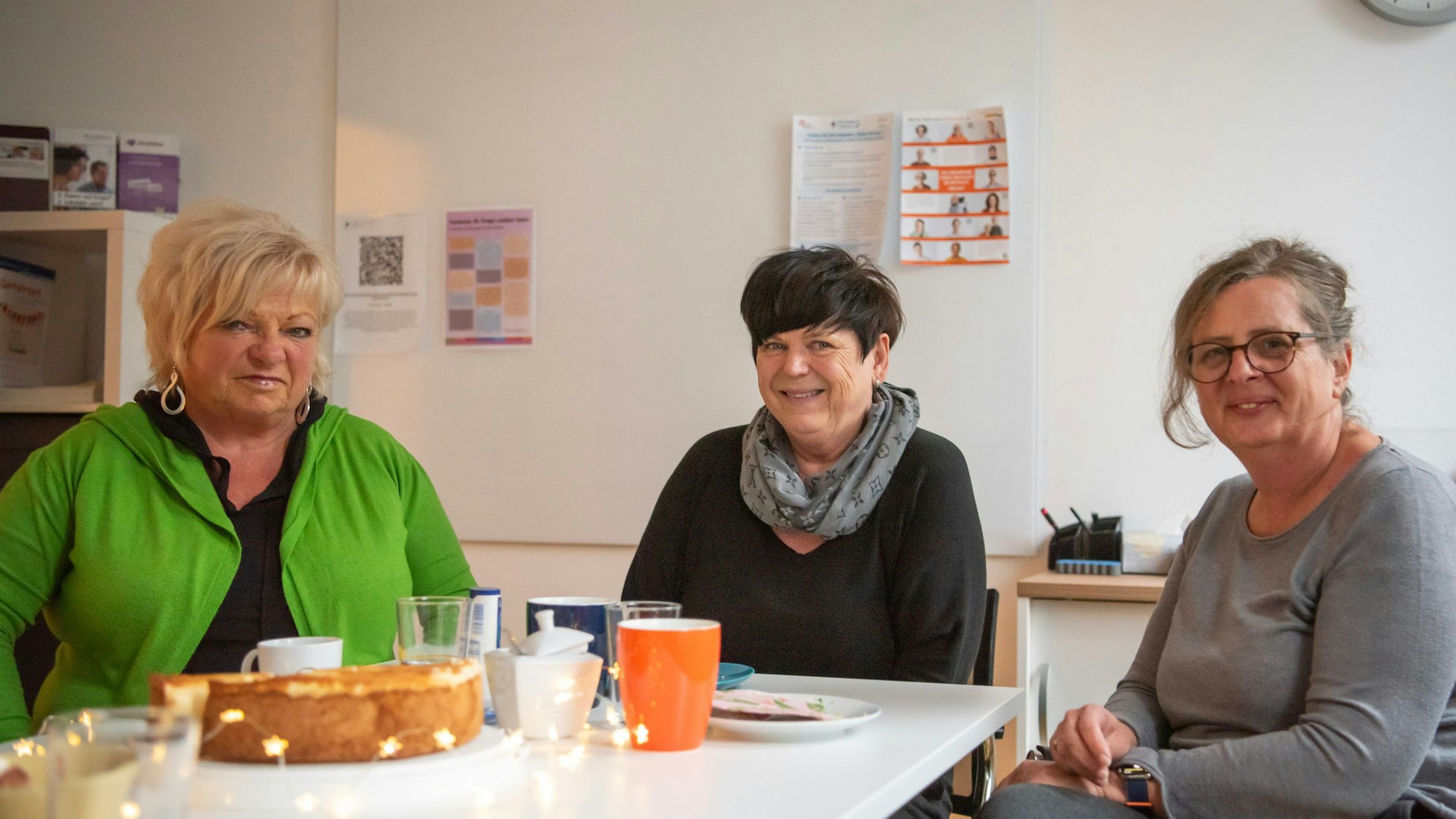 Die drei Frauen sitzen an einem Tisch in einem Büroraum. Auf dem Tisch stehen ein Käsekuchen und Kaffeegeschirr, daneben liegt eine Sternen-Lichterkette.