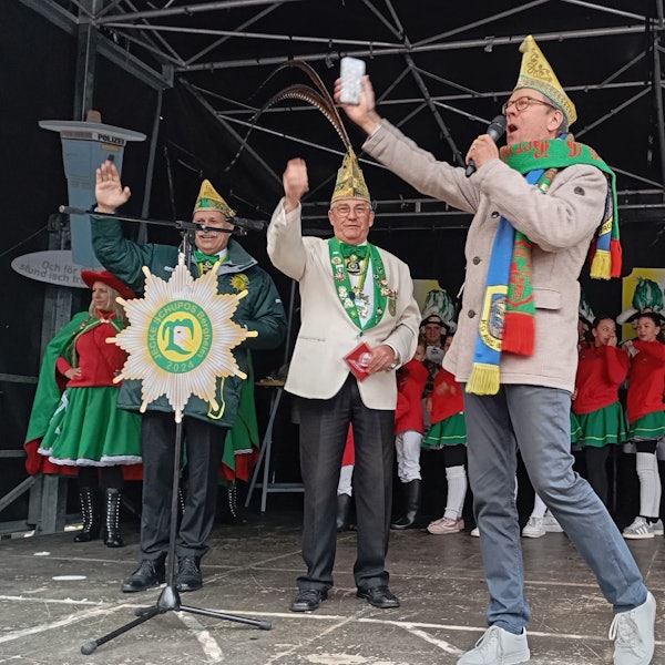 Drei Männer mit Narrenkappen stehen auf einer Bühne.