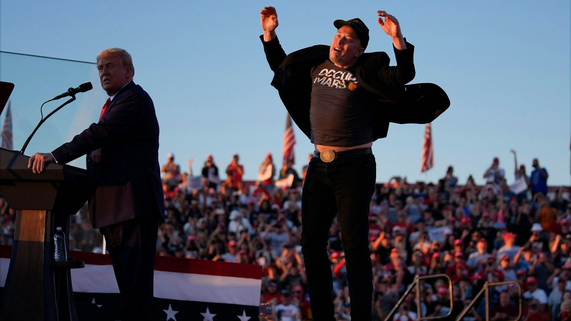 ARCHIV - 05.10.2024, USA, Butler: Elon Musk springt auf der Bühne, während der republikanische Präsidentschaftskandidat und ehemalige US-Präsident Donald Trump bei einer Wahlkampfveranstaltung bei der Butler Farm Show spricht.