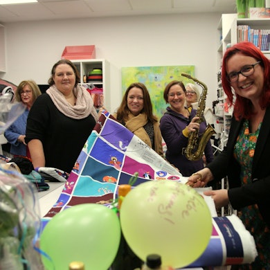 Frauen lächeln in die Kamera, auf einem Tisch vor ihnen liegen Luftballons und eine Stoffbahn.