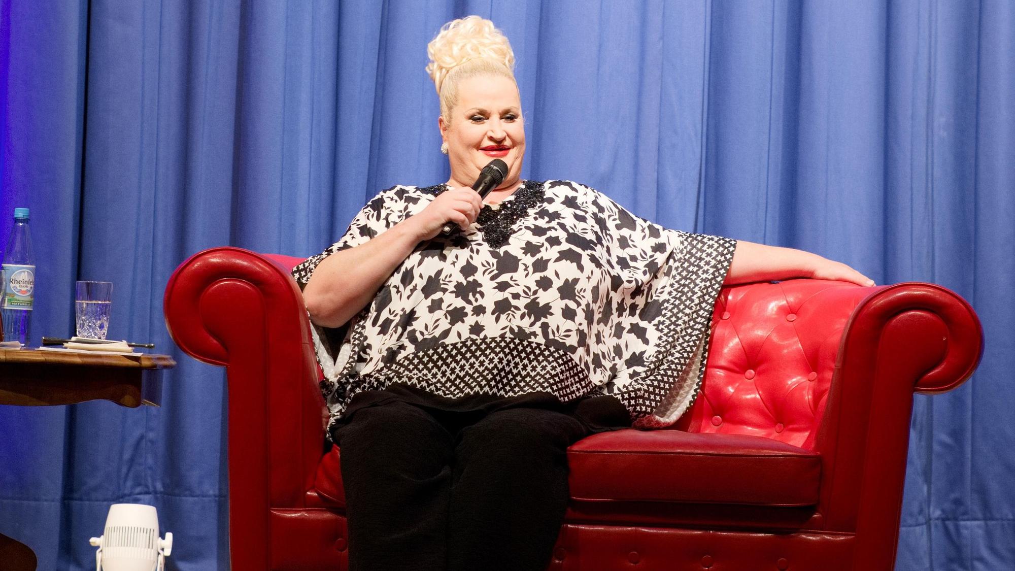 Daphne de Luxe sitzt auf einer roten Couch im Euskirchener Stadttheater und spricht in ein Mikro.