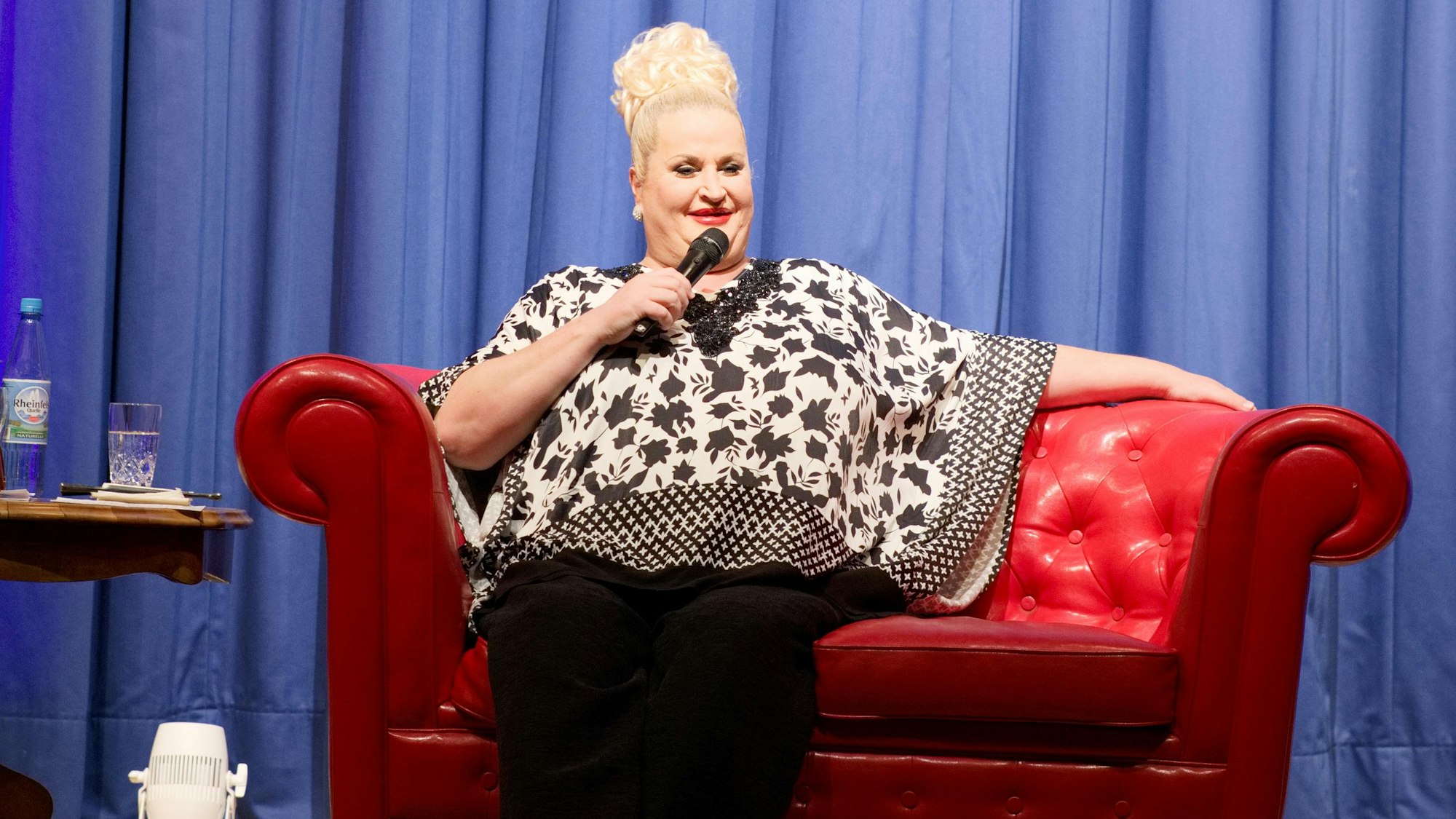 Daphne de Luxe sitzt auf einer roten Couch im Euskirchener Stadttheater und spricht in ein Mikro.