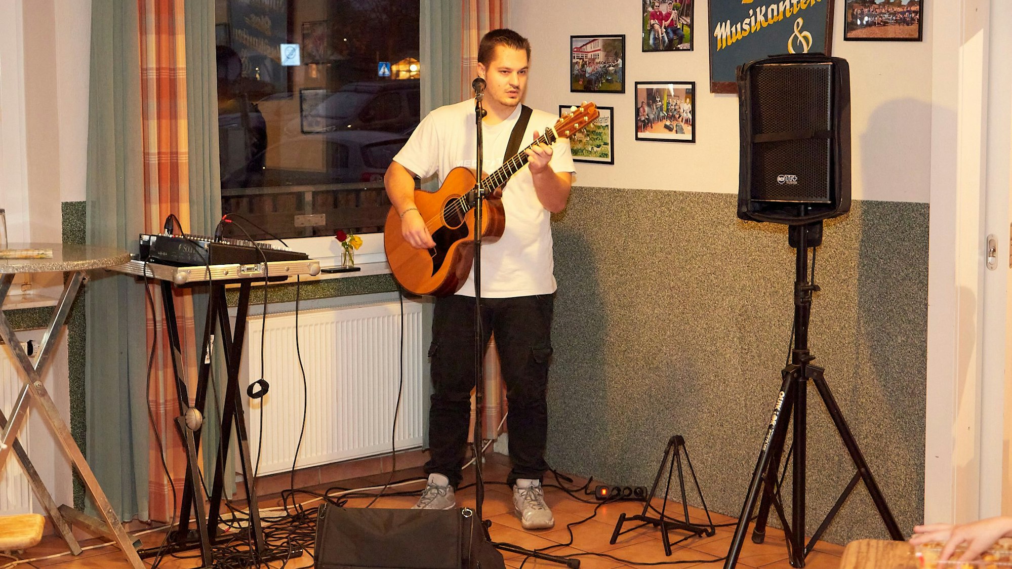 Ein junger Mann in weißem T-Shirt und schwarzer Jeans steht mit einer Gitarre an einem Mikrofon.