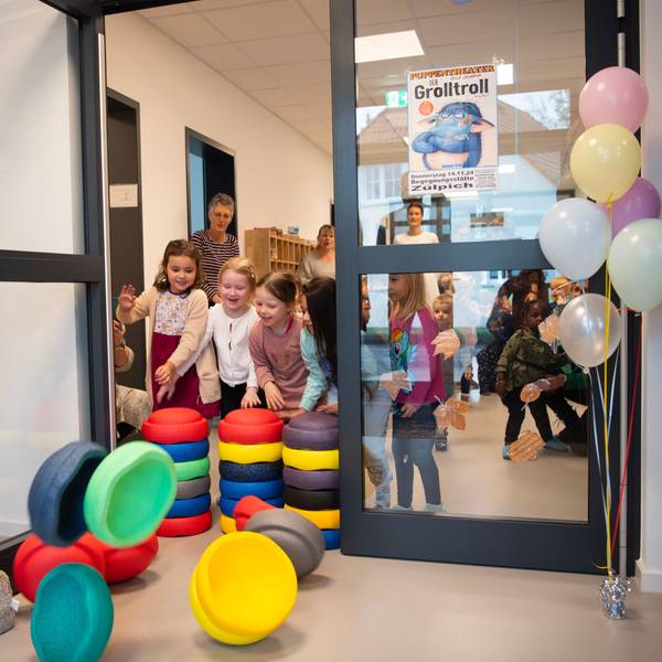 Kinder schmeißen bunte Stapelsteine, die eine Tür versperren, um. Neben der Tür steht ein Strauß bunter Luftballons.