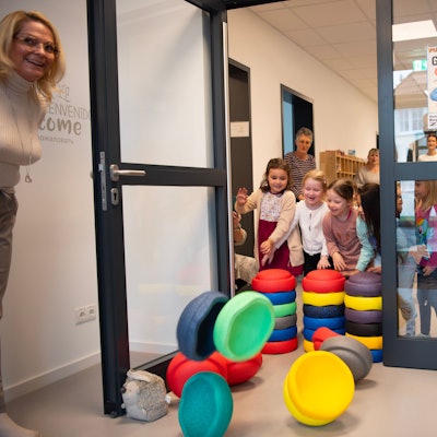 Kinder schmeißen bunte Stapelsteine, die eine Tür versperren, um. Neben der Tür steht ein Strauß bunter Luftballons.