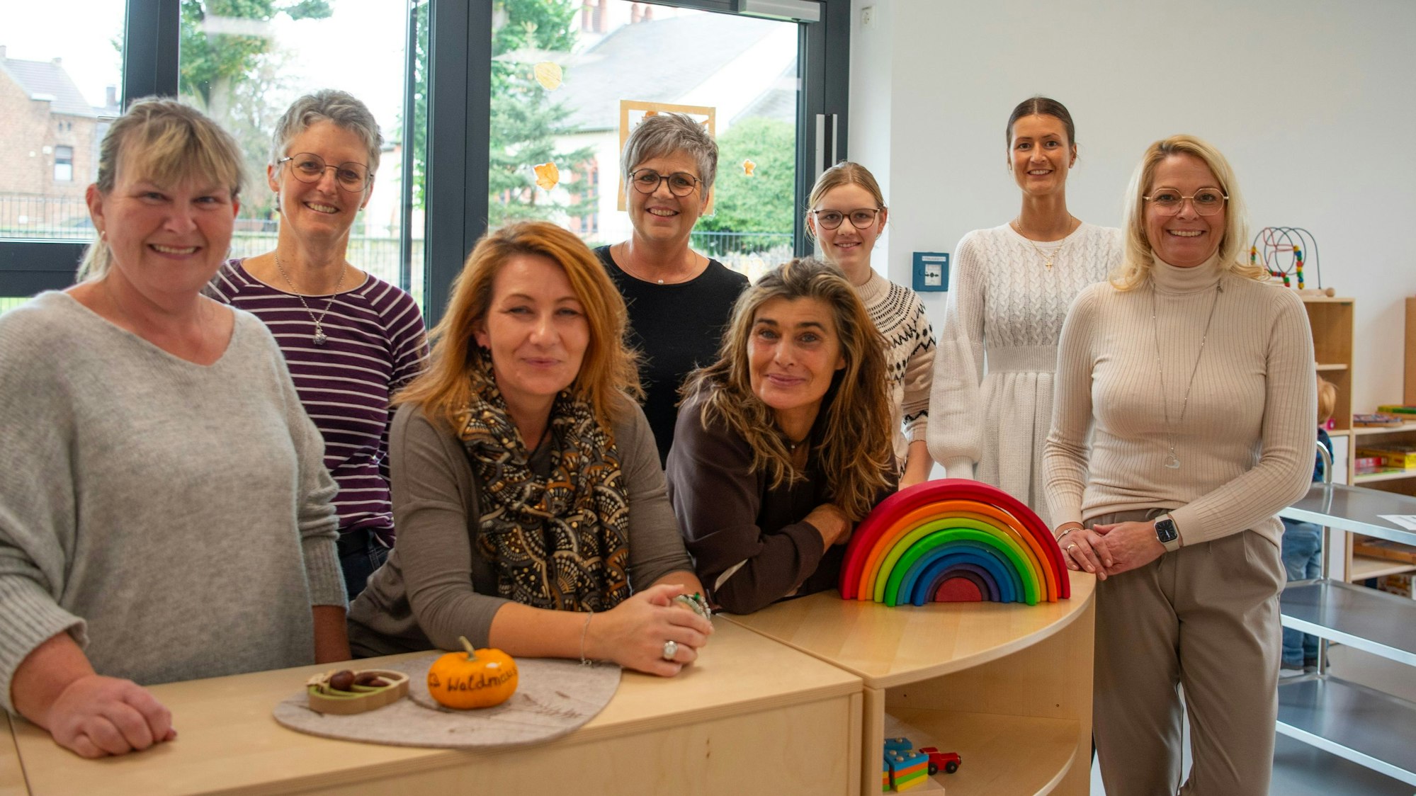 Das Team der Kita Springmäuse steht an einem neuen Spieleregal, im Hintergrund sind große Fenster. Auf dem Regal liegen ein bunter Regenbogen und ein Kürbis mit der Aufschrift „Waldmäuse“.