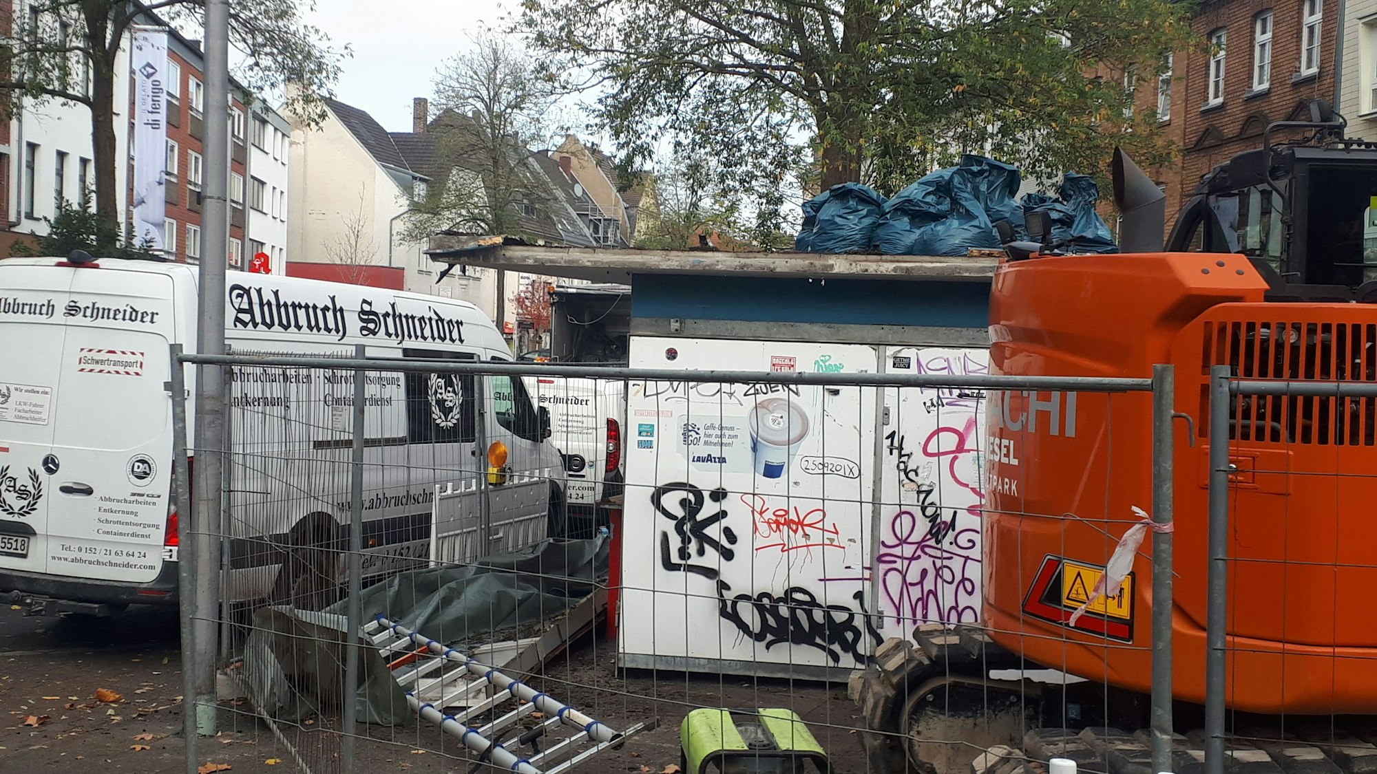 Hinter einem Bauzaun steht schweres Gerät und ein kleines flaches Gebäude.