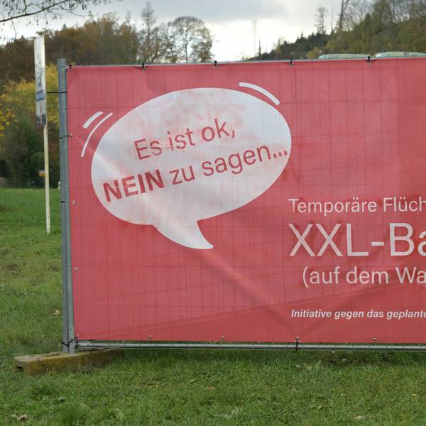 Ein großes, rotes Plakat steht auf einer Wiese. Darauf ist in einer Sprechblase zu lesen: "Es ist okay, NEIN zu sagen...". Und: "Temporäre Flüchtlingsunterkunft JA - XXL-Bau NEIN". Initiative gegen das geplante Bauvorhaben in Odenthal-Höffe.
