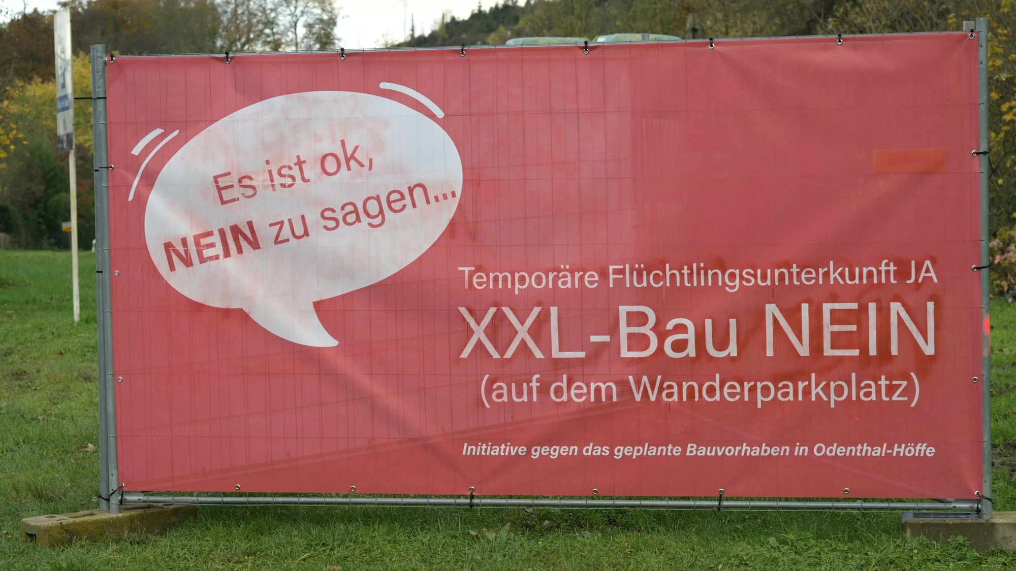 Ein rotes Plakat steht an einer Straße. Darauf der Protest einer Bürgerinitiative gegen den Bau eines Flüchtlingsheims in Odenthal.