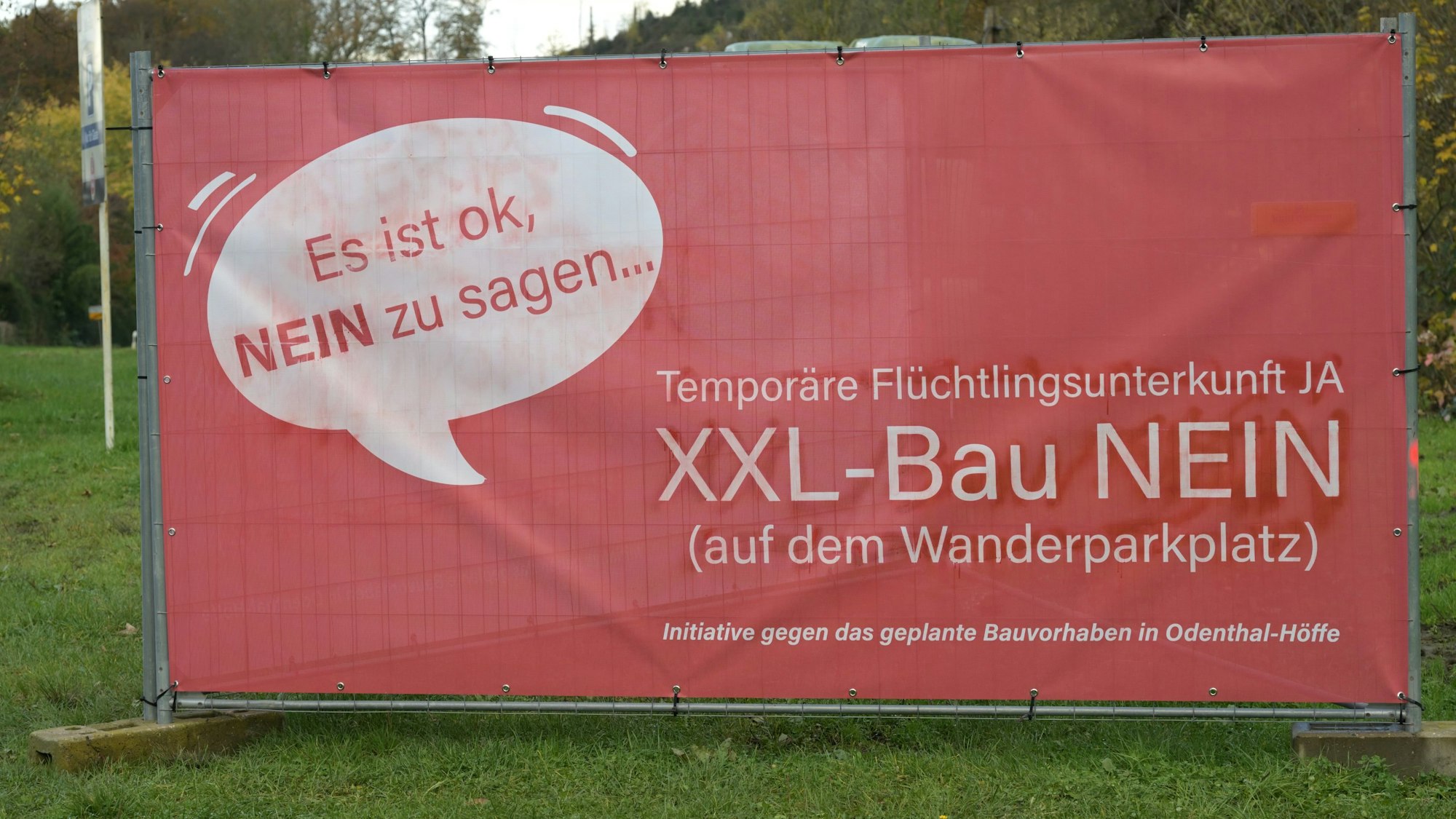 Ein großes, rotes Plakat steht auf einer Wiese. Darauf ist in einer Sprechblase zu lesen: "Es ist okay, NEIN zu sagen...". Und: "Temporäre Flüchtlingsunterkunft JA - XXL-Bau NEIN". Initiative gegen das geplante Bauvorhaben in Odenthal-Höffe.