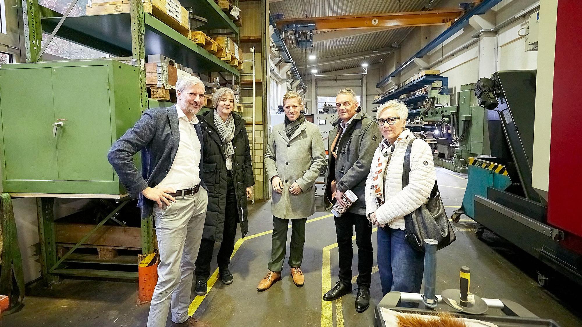 Eine Gruppe Menschen steht in einer großen Werkshalle. Links ist ein hohes grünes Regal zu sehen, in dem Paletten lagern.