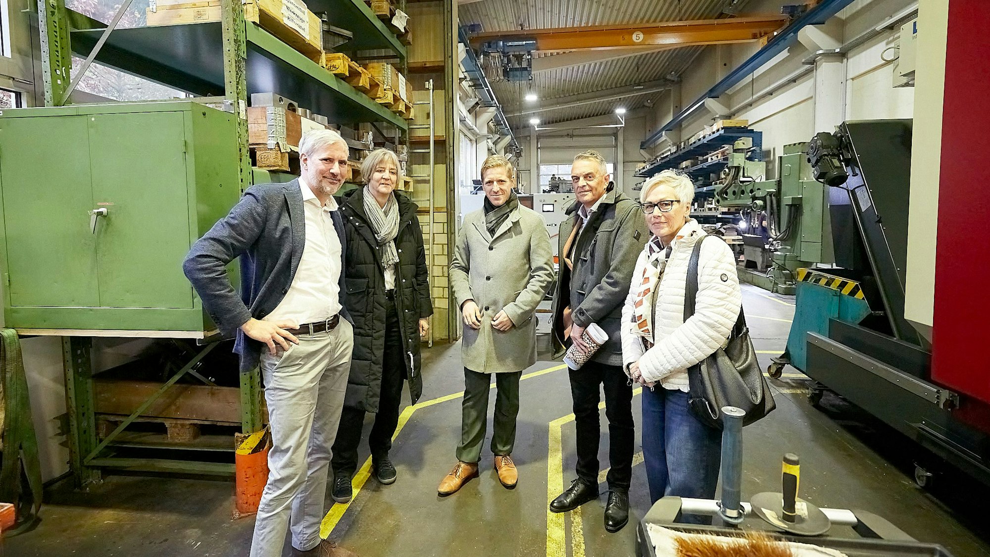 Eine Gruppe Menschen steht in einer großen Werkshalle. Links ist ein hohes grünes Regal zu sehen, in dem Paletten lagern.