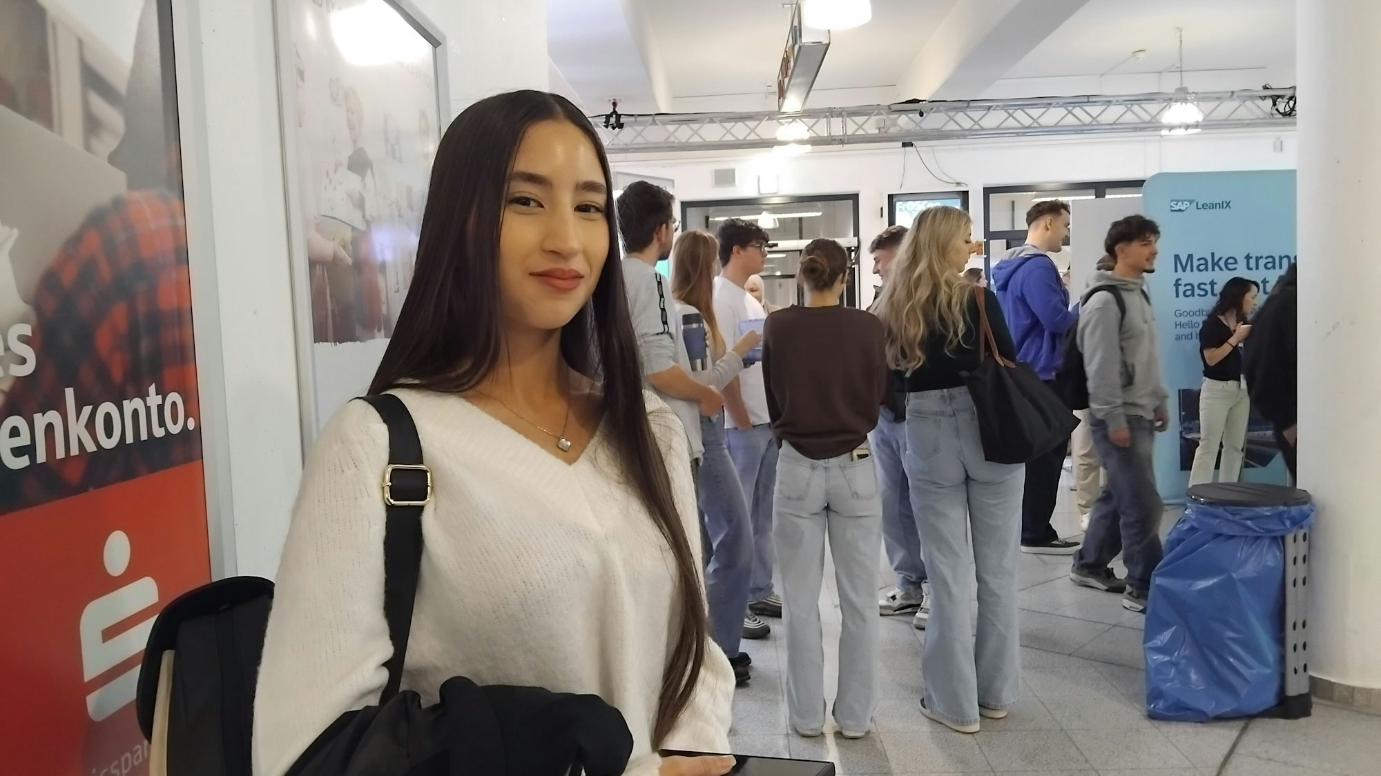 Eine Studentin steht in einem Raum, wo man im Hintergrund Aufstellplakate und viele weitere Studierende sieht.