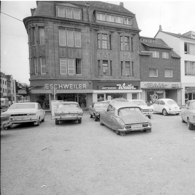 Auf einem Platz stehen Autos, im Hintergrund die Neustraße mit Geschäftslokalen.