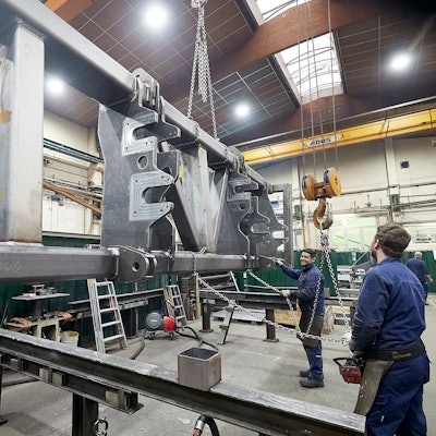 Zwei Männer in blauer Arbeitskleidung stehen in einer großen Halle und bedienen einen Kran, an dem ein großes Stahlteil hängt.