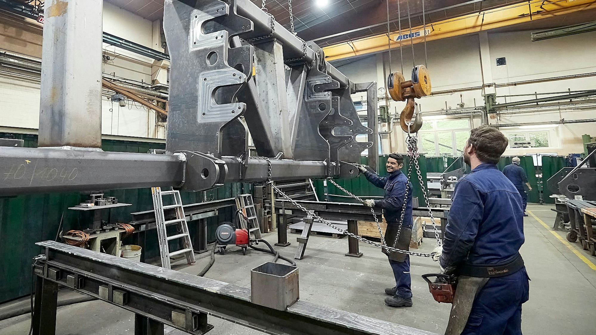 Zwei Männer in blauer Arbeitskleidung stehen in einer großen Halle und bedienen einen Kran, an dem ein großes Stahlteil hängt.