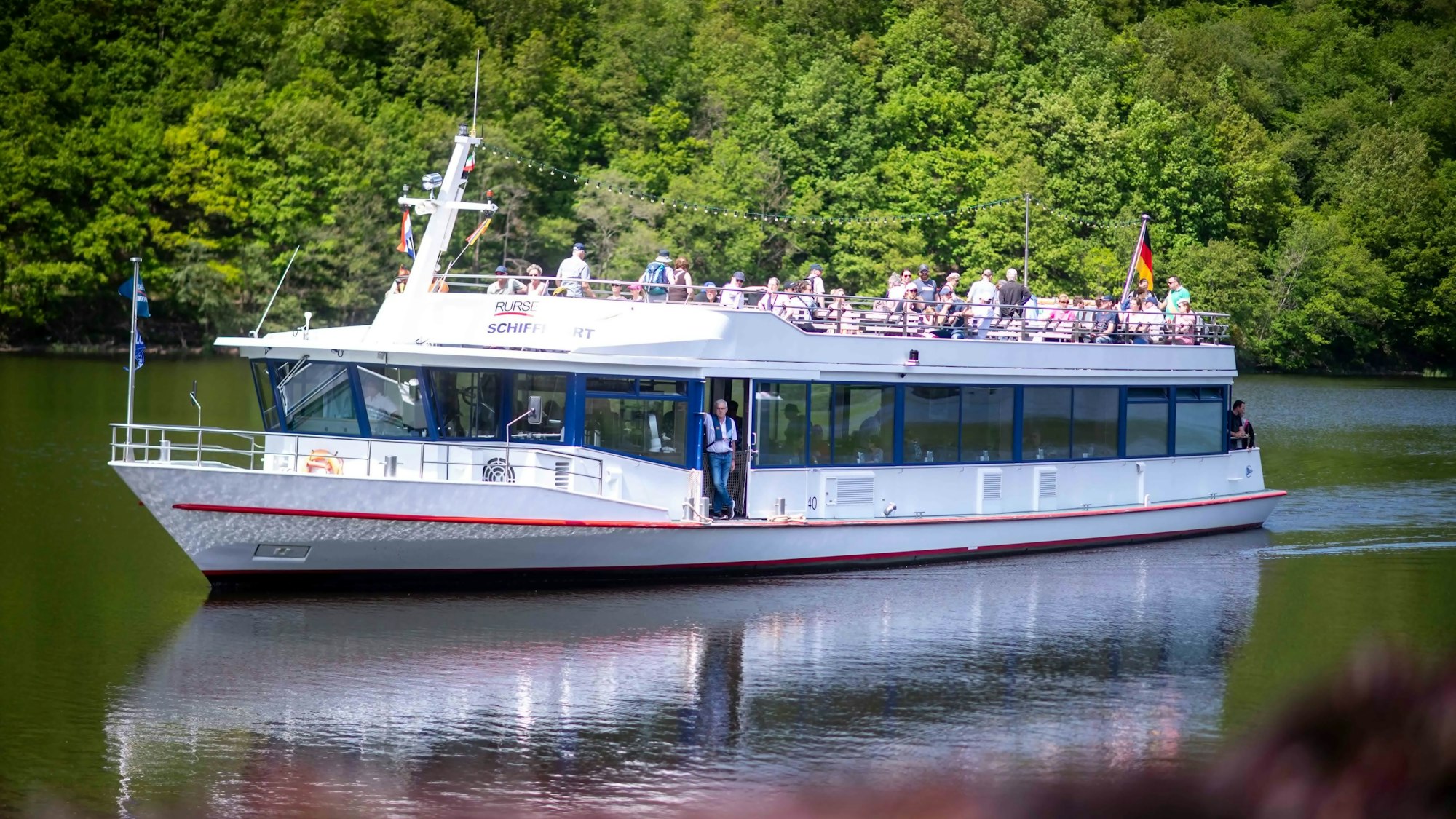 Ein weißes Schiff fährt auf dem Rursee, im Hintergrund sieht man grünen Wald.